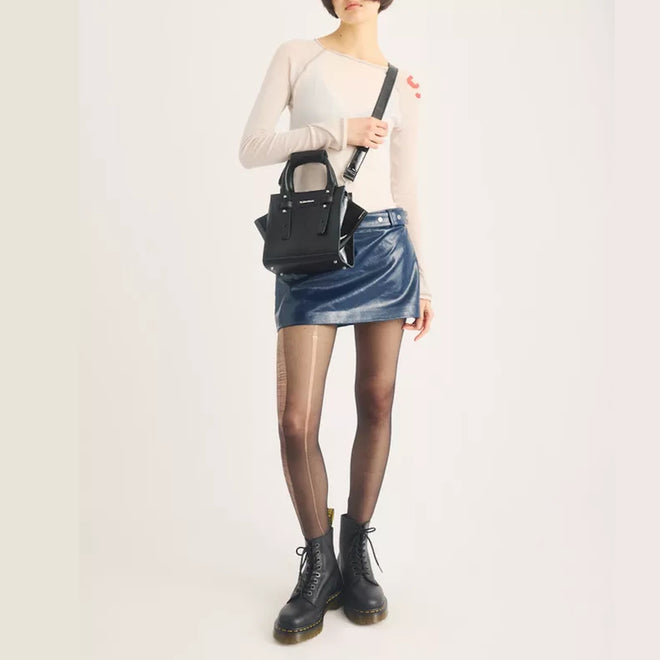 A person wearing a fitted white top, blue mini skirt, sheer tights, and black lace-up boots crafted from sleek black Kiev leather stands against a plain light background, carrying the Dr Martens Small Keiv Leather Tote Bag Black over their shoulder.