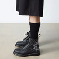 A person in a black skirt and socks wears Dr Martens - 1460 Genix Solar Flare Black 8 Up Eye Boots, featuring a white sunburst design, standing on a light-colored floor against a plain background., 190665742824, 190665742831, 190665742848, 190665742862, 190665742879, 190665742886