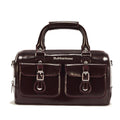 A glossy dark brown handbag inspired by the Dr Martens handle bag design, featuring two front pockets, buckle details, silver hardware, and a sturdy top handle. The brand name is visible above the pockets.