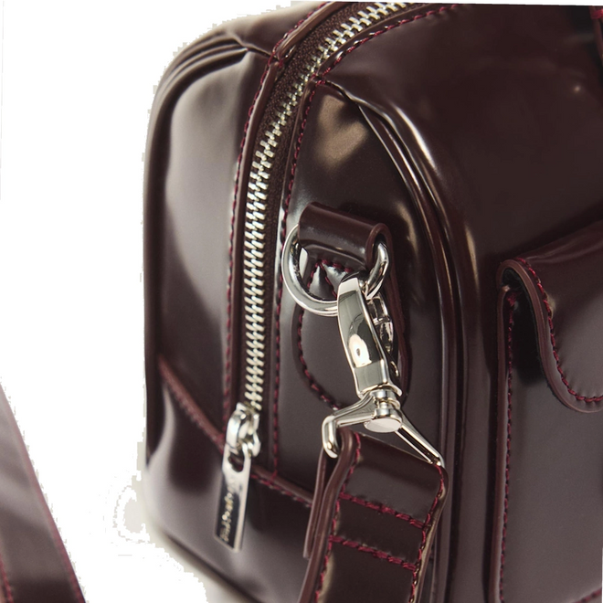 Close-up of the Dr Martens Top Handle Bag in Cherry Red Oxford Rub Off, featuring glossy burgundy leather with silver hardware, including a zipper, detailed stitching, and a metal clasp on the strap—an elegant accessory showcasing the signature oxford rub off finish.