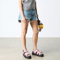 A person in a gray and white striped tank top, ripped denim shorts, and Dr Martens Voss II Cross Strap Sandal Dusty Rose Pink stands indoors holding a small yellow-and-black bag. The photo is cropped at the shoulders., 50439, 190665777529, 190665777536, 190665777543, 190665777567, 50444, 50445