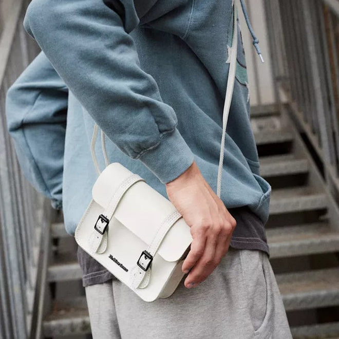 A person in a blue sweatshirt and gray pants stands on stairs, wearing the Dr Martens Leather Cross Body White Leather Box Satchel—a stylish small white leather bag that completes the look., 190665390384