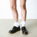 A person showcases Dr Martens Ankle Frill Cotton Blend Socks White with black lace-up shoes, standing on a light floor against a white wall. Only their legs and feet are visible, drawing attention to the frilly ankle socks' charming design., 190665640854, 190665640847