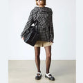 A person wearing a black-and-white checkered oversized top, tan shorts, black sheer tights, and Dr Martens Women's Mary Jane BW Black + White shoes stands against a plain background, carrying a large black handbag., 190665785333, 190665785340, 190665785364, 50336, 50337