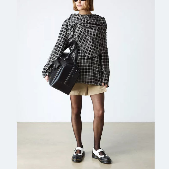 A person wearing a black-and-white checkered oversized top, tan shorts, black sheer tights, and Dr Martens Women's Mary Jane BW Black + White shoes stands against a plain background, carrying a large black handbag., 190665785333, 190665785340, 190665785364, 50336, 50337