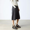 A person wearing a long-sleeve cream top, dark knee-length wide shorts, and Dr Martens Gryphon Sandal Black Atlas Leather stands indoors on a light gray floor, facing slightly to the side against a plain white background., 190665783414, 190665783421, 190665783438, 190665783452, 190665783469, 50536