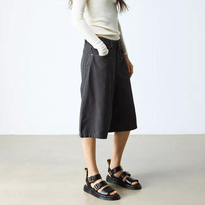 A person wearing a long-sleeve cream top, dark knee-length wide shorts, and Dr Martens Gryphon Sandal Black Atlas Leather stands indoors on a light gray floor, facing slightly to the side against a plain white background., 190665783414, 190665783421, 190665783438, 190665783452, 190665783469, 50536