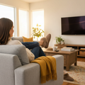 A person wearing EMU Womens Mintaro Sheepskin Slipper in Mushroom relaxes on a gray sofa with a yellow blanket, watching a wall-mounted TV in a bright, cozy living room filled with plants and books., 51402, 9330071981665, 9330071981672, 9330071981689, 51406, 51407, 51408