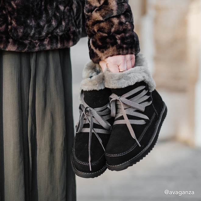 A person in a furry animal-print jacket and pleated pants holds Emu Womens Thorsby Black Sheepskin Ankle Boot Waterproof, featuring gray laces, arch support, and faux fur lining. The neutral background is slightly blurred., 9330071980507, 9330071980514, 9330071980521