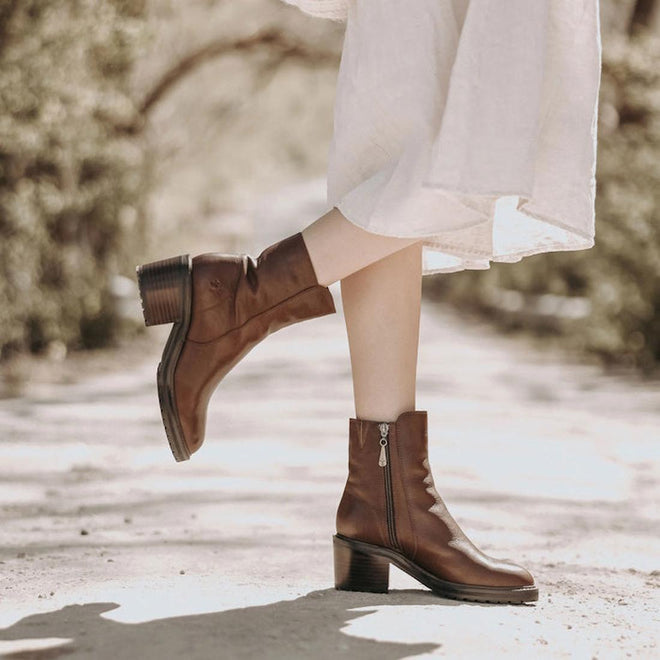 A model wearing a pair of EMU womens brown leather EMU Zeil ankle boots with a medium height block heel. The right foot is raised showing the outer side of the boot and the left foot is on the ground showing the inner zip., 9330071964217,
9330071964224,
9330071964231,
9330071964248,