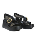 The EOS Arianis Black Womens Leather Platform Wedge Sandals, crafted from black leather with crisscross straps and gold buckle accents, feature an anatomical footbed and an adjustable ankle strap, elegantly displayed against a white background.