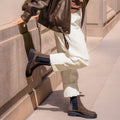 A person in light pants, a brown leather jacket, and EOS Womens Leilin Suede Kalamata Brown Ankle Pull On Boots stands on the sidewalk, one foot raised and resting against a stone building wall., 9321190037555, 9321190037562, 9321190037579, 9321190037586, 9321190037593, 50608, 50609