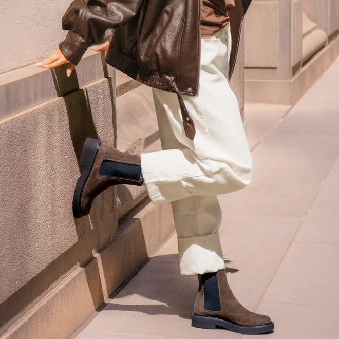 A person in light pants, a brown leather jacket, and EOS Womens Leilin Suede Kalamata Brown Ankle Pull On Boots stands on the sidewalk, one foot raised and resting against a stone building wall., 9321190037555, 9321190037562, 9321190037579, 9321190037586, 9321190037593, 50608, 50609