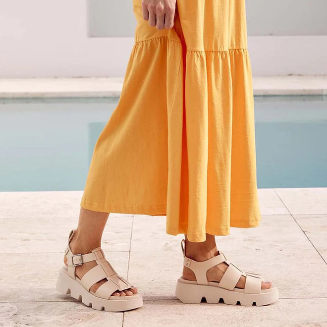 A person wearing a long yellow dress and EOS Womens Kailan Platform Chunky Leather Sandals in Bone stands on a light stone surface near a pool with calm blue water., 34434, 9321191131504, 9321191131511, 9321191131528, 9321191131535, 9321191131542, 9321191131559