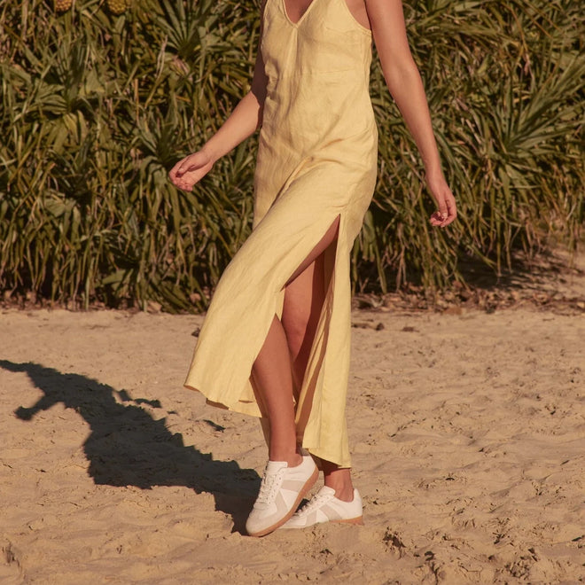 A person in a sleeveless yellow dress with a high slit walks barefoot on sand, holding EOS Melbaa White Leather Lace Up Trainers, with green plants behind them. Their head is not visible., 41691, 9321191224626, 9321191224633, 9321191224640, 9321191224657, 9321191224664, 41697