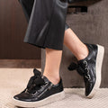 A person wearing black leather pants and EOS Joyce Leather Trainer Black Lace Up with Zip sneakers, featuring thick soles, large black laces, anatomical insoles, and side zippers, rests their feet on an indoor concrete surface.