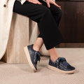 A person sits on a bench, wearing black pants, a light coat, and EOS Jovi Leather Trainer Navy Lace Up with Zip—comfortable sneakers made from Italian Aniline leather, with thick beige soles and bold blue laces. The floor is carpeted in light color.