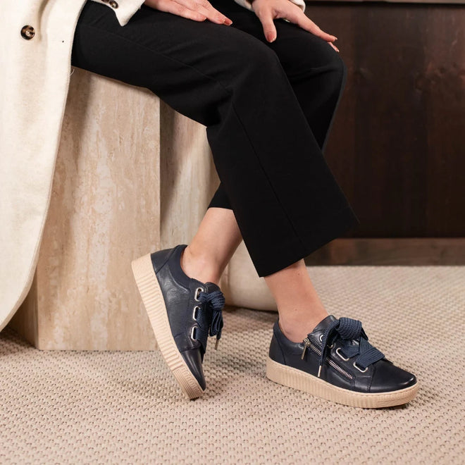 A person sits on a bench, wearing black pants, a light coat, and EOS Jovi Leather Trainer Navy Lace Up with Zip—comfortable sneakers made from Italian Aniline leather, with thick beige soles and bold blue laces. The floor is carpeted in light color.