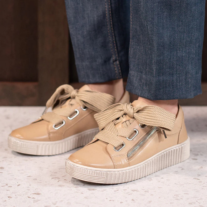 Wearing blue jeans, a person stands on a speckled white floor in EOS Jovi Leather Trainer Tan sneakers featuring thick laces and metallic side zippers for both comfort and style.
