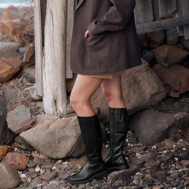 A person stands on rocky ground, wearing EOS Blanche Womens Black Leather High Boot Low Heel with a dark brown coat and tan skirt. Large rocks and wooden beams are in the background. The head and upper body are not visible., 9321191187808, 9321191187815, 9321191187822, 9321191187839, 9321191187846, 50595