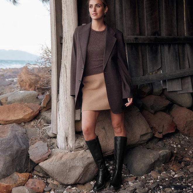 A woman stands outdoors by a rustic wooden structure, wearing the EOS Wyoming Black High Leather Women’s Heeled Boot, along with a brown blazer, brown sweater, and tan skirt. Rocky ground and distant water are seen in the background., 9321191201733, 9321191201740, 9321191201757, 9321191201764, 9321191201771, 9321191201788