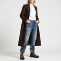 A person wearing EOS Womens Leilin Suede Kalamata Brown Ankle Pull On Boots, a long brown coat, white button-up shirt, and blue embroidered jeans stands with hands in jean pockets against a plain white background., 9321190037555, 9321190037562, 9321190037579, 9321190037586, 9321190037593, 50608, 50609