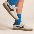 A person wearing bright blue socks and Gelato Staton Swan Off White / Cafe Leather Lace Womens Sneakers stands on one foot, lifting the other leg off the ground against a light background., 52483, 52484, 52485, 52486, 52487, 52488