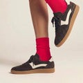A person wearing Gelato Staton Chocolate Brown / Swan Leather Lace Womens Sneakers with bright red socks stands on one foot, the other slightly raised, set against a neutral background., 52477, 52478, 52479, 52480, 52481, 52482