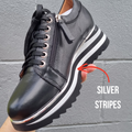 A hand holds the Alfie & Evie Hosting Womens Leather Shoe Platform Black Silver, a lightweight leather sneaker with a side zipper and thick sole, featuring shiny silver stripes on the midsole. An arrow labeled SILVER STRIPES highlights this detail., 50827, 50828, 50829, 50830, 50831, 50832, 50833