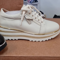 The Alfie & Evie Hosting Womens Leather Shoe in Cream/Gold, featuring a thick platform sole and gold accents, is displayed atop a brown box, with other footwear softly blurred in the background.