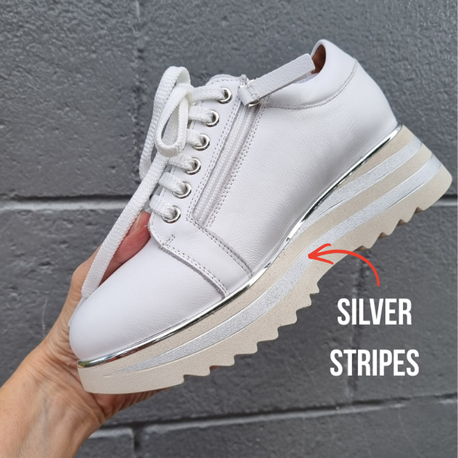 A hand holds the Alfie & Evie Hosting Womens Leather Shoe Platform White/Silver, featuring white laces, a zipper, and silver metallic stripes on the midsole. "SILVER STRIPES" text with an arrow highlights the detail against a gray brick wall.