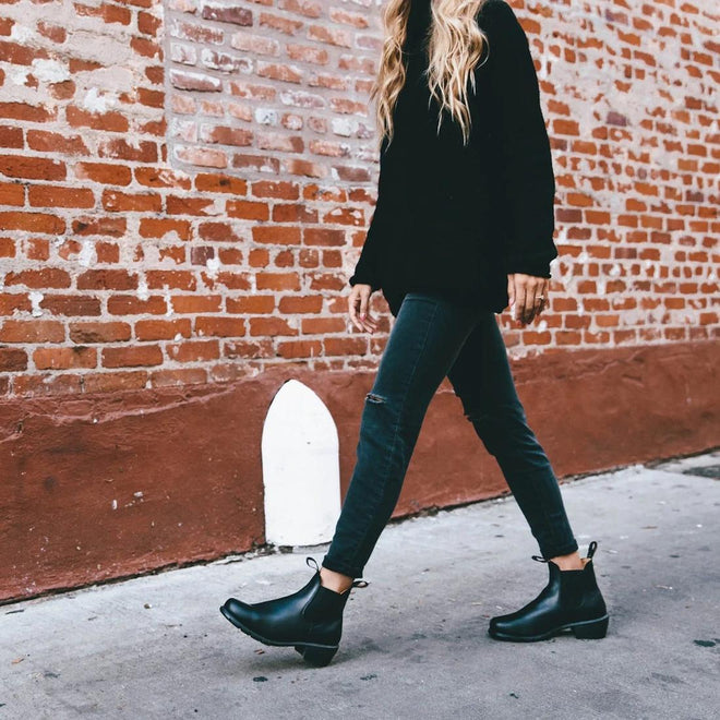 A person with long, wavy blonde hair walks by a brick wall in a black sweater, dark jeans, and Blundstone Womens 1671 Low Heeled Black Ankle Chelsea Boots., 10445, 9315891491846, 9315891491860, 9315891491884, 9315891491907, 9315891491921, 9315891491945