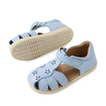 Introducing the Bobux IWalk Holly Toddlers Sandal in Skyway Blue: closed toe sandals for children with delightful cut-out flower motifs, a secure Velcro ankle strap, and beige rubber soles. Made from quick-drying leather, these sandals provide both comfort and long-lasting wear for toddlers. In this image, one sandal stands upright while the other showcases its sole.