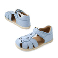 A pair of Bobux IWalk Holly Toddlers Sandals in Skyway Blue, featuring closed toes, quick-drying leather, charming cutout flower designs, and beige soles. One sandal is angled to the left while the other faces forward.