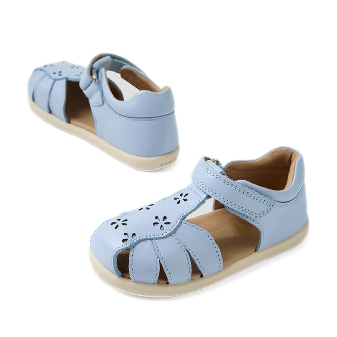 A pair of Bobux IWalk Holly Toddlers Sandals in Skyway Blue, featuring closed toes, quick-drying leather, charming cutout flower designs, and beige soles. One sandal is angled to the left while the other faces forward.