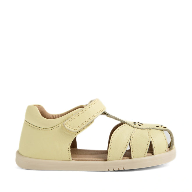 The Bobux IWalk Holly Toddlers Sandal in Wax Yellow is a closed-toe sandal for little explorers, crafted from quick-drying leather with a pale yellow finish. It features a closed heel, open sides, Velcro strap, and stylish cut-out details on the toe area—shown here against a white background.