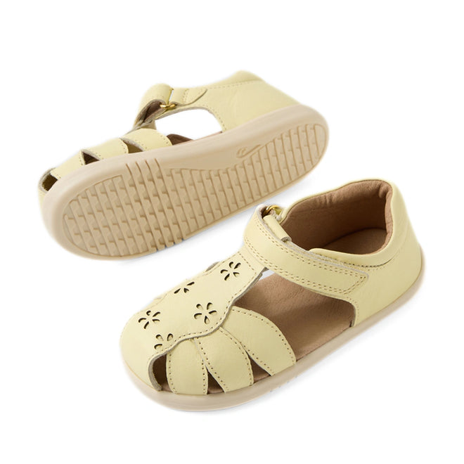 The Bobux IWalk Holly Toddlers Sandal in Wax Yellow features a light yellow closed-toe design for toddlers, crafted from quick-drying leather with delightful cut-out flower details. These sandals include a Velcro strap and flexible beige rubber soles. One shoe is positioned upright while the other showcases its sole.