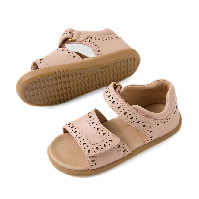 Two Bobux IWalk Logan Toddlers Sandals in Cameo Rose Pink, featuring an open toe, perforated decorative edges, tan soles, and hook-and-loop straps. One leather sandal is upright, while the other displays its textured sole.