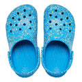 The Crocs Kids - Toddlers Classic Clog K Paint Splatter Print Blue Pink Multi features lightweight blue clogs for kids with colorful paint splatters, ventilation holes, and adjustable heel straps, shown from above on a white background., 47582, 198445214134, 198445214141, 198445214158, 198445214097