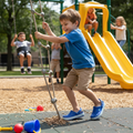 A young boy in a blue shirt and shorts, wearing Crocs Kids - Crocband Classic Clog K Blue Bolt Teal Youths, climbs a rope ladder at the playground while other children play on swings and a yellow slide among colorful toys on a sunny day., 49846, 198445353406, 198445353413, 198445353420, 198445353437, 198445353444, 198445353451, 49853