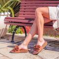 A person sits outdoors on a maroon bench, wearing light shorts and Los Cabos Corsage Truffle Brown Flower Women's Slides with wedge heels and large fabric flowers. Nearby are a large potted plant and stone pavement., 9321191299174, 9321191299181, 9321191299198, 9321191299204, 9321191299211, 46759