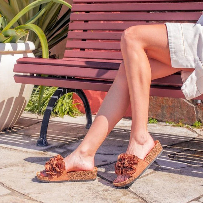 A person sits outdoors on a maroon bench, wearing light shorts and Los Cabos Corsage Truffle Brown Flower Women's Slides with wedge heels and large fabric flowers. Nearby are a large potted plant and stone pavement., 9321191299174, 9321191299181, 9321191299198, 9321191299204, 9321191299211, 46759