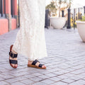 A person in a long white lace dress walks on a paved sidewalk lined with large planters, wearing Los Cabos Doti Black Light Flexible-Sandals., 9321191045450, 9321191045467, 9321191045474, 9321191045481, 9321191045498, 9321191045504, 9321191045511