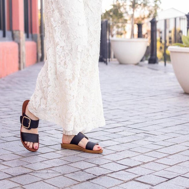 A person in a long white lace dress walks on a paved sidewalk lined with large planters, wearing Los Cabos Doti Black Light Flexible-Sandals., 9321191045450, 9321191045467, 9321191045474, 9321191045481, 9321191045498, 9321191045504, 9321191045511