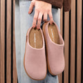 A person in blue jeans holds a pair of Los Cabos Felton Graphic Lavender Women's Felted Slipper House slippers, featuring pink felt and cushioned brown soles, against a backdrop of vertical wooden panels., 9321190006025, 9321190006032, 9321190006049, 9321190006056, 9321190006063, 9321190006070, 50087