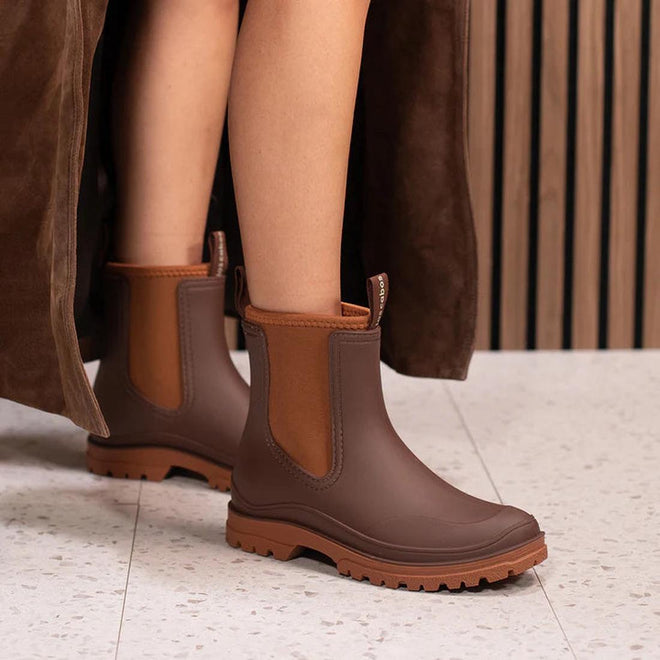 Close-up of a person wearing Los Cabos Amelia Chocolate Ankle Rain Boot Waterproof, featuring rugged soles and a neoprene lining, on a light speckled floor. Part of a brown coat and wooden wall appear in the background., 50781, 9321190004915, 9321190004922, 9321190004939, 9321190004946, 9321190004953, 9321190004960