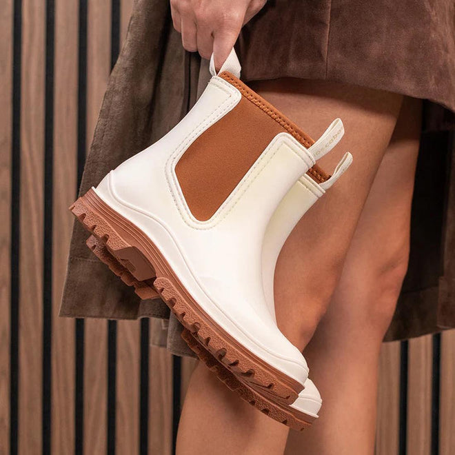 A person in a brown skirt holds the Los Cabos Amelia Cream Ankle Rain Boot Waterproof—trendy chunky Chelsea-style rain boots with rugged soles—standing before a vertical wooden slat wall., 9321190004977, 9321190004984, 9321190004991, 9321190005004, 9321190005011, 9321190005028, 50794