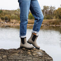 A person in rolled-up blue jeans and Los Cabos Amelia Khaki Ankle Rain Boot Waterproof stands on a rock by a calm lake, with trees and shrubs in the background., 50795, 9321191166711, 9321191166728, 9321191166735, 9321191166742, 9321191166759, 9321191166766