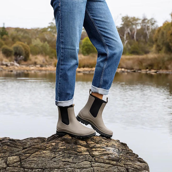 A person in rolled-up blue jeans and Los Cabos Amelia Khaki Ankle Rain Boot Waterproof stands on a rock by a calm lake, with trees and shrubs in the background., 50795, 9321191166711, 9321191166728, 9321191166735, 9321191166742, 9321191166759, 9321191166766