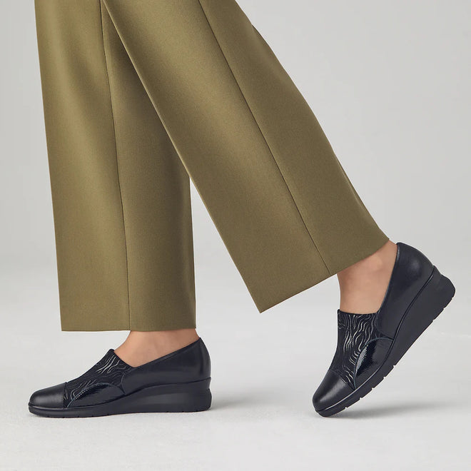 A person stands on a light gray floor wearing olive green pants and Pitillos Womens Lycra and Patent Leather Shoe 10731, featuring a black wedge heel, subtle textured pattern, and removable innersole, against a plain background., 8445191655554, 8445191655561, 8445191655578, 8445191655585, 51923, 51924